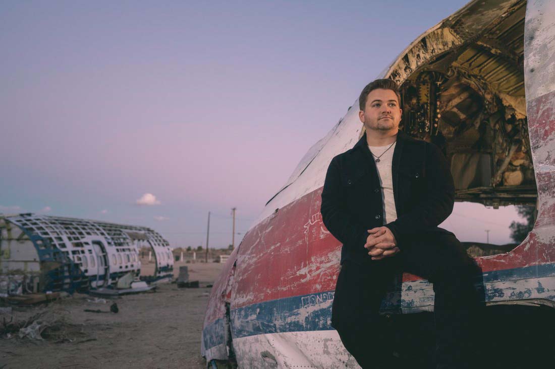 Hunter Hayes - Photo by Andrw Zaeh. Hunter sits in the doorway of a dilapidated airplane as a rosy sunset frames the sky.