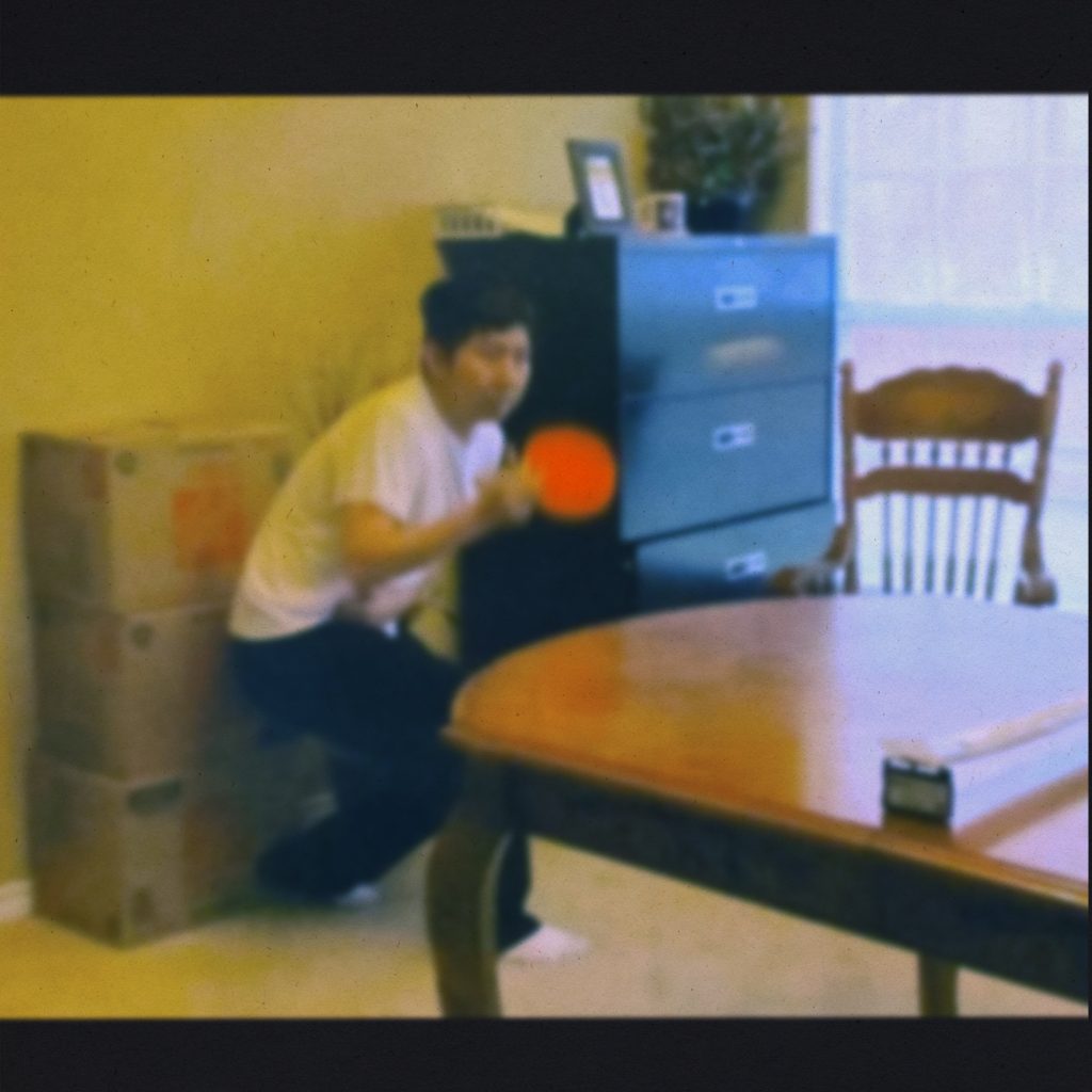 Kaz Moon - "A lot to explain" - album art - a blurry image of Kaz playing ping pong on a dining room table while inside a home