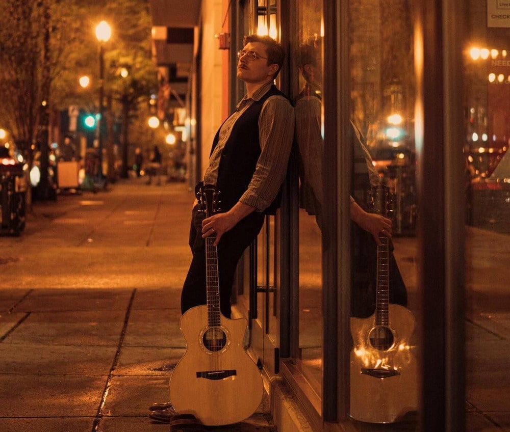 Eli with guitar leaning on building window at nighttime