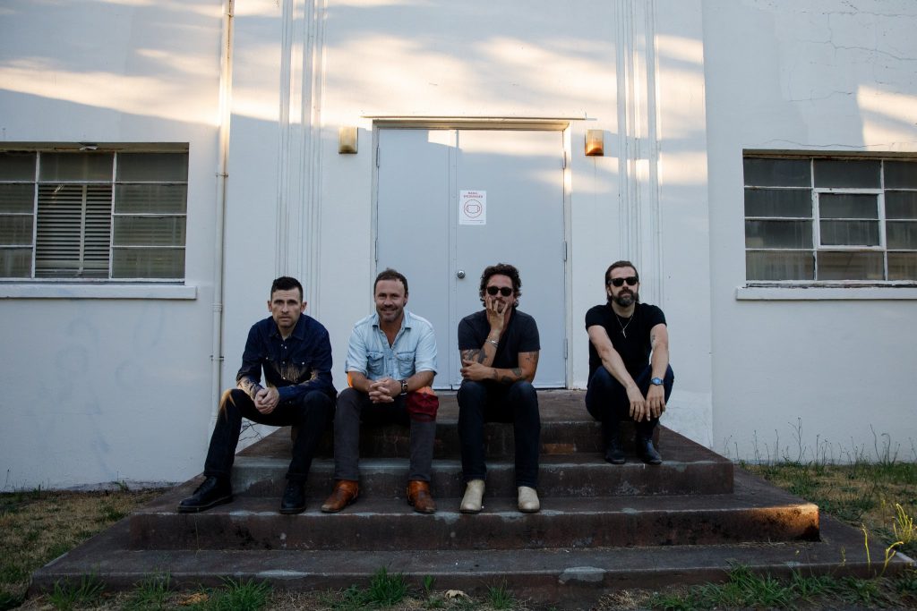Four bandmates sit on a small set of stairs front of a door to a big white building.
