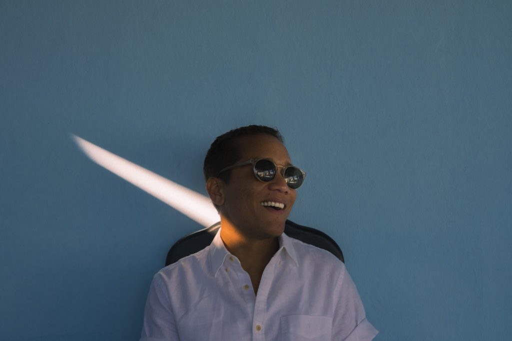 Man laughs while looking away away from the camera wearing a white shirt and sunglasses while sitting in a black chair against a blue wall with a sunbeam streaked across it.