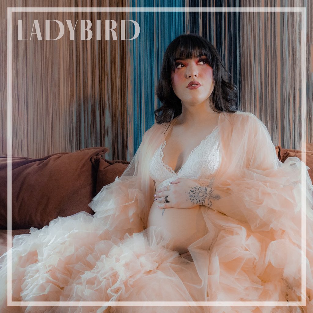 Woman in white lingerie sits on a brown couch in front of a multi-colored background while she looks away from the camera with her hand resting on her belly.