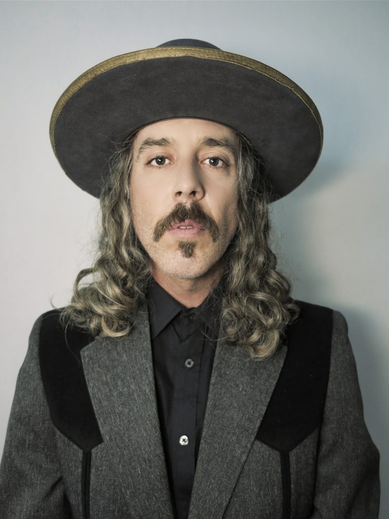 Mustached, long haired man looks into the camera wearing a big rimmed hat, gray blazer, and a button up shirt.
