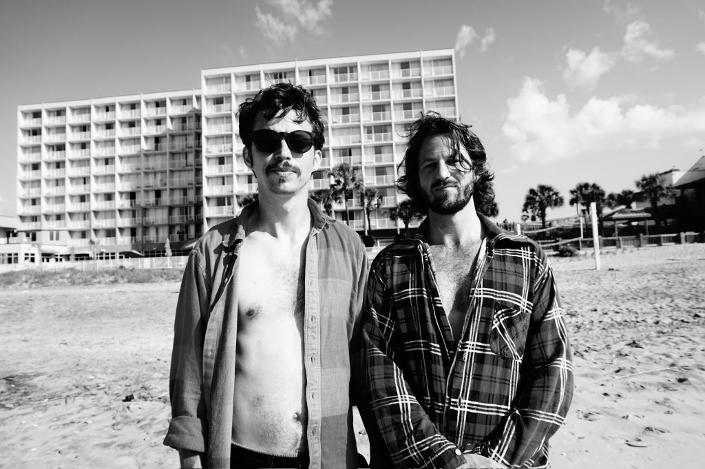 Two men wearing loosely buttoned shirts and disheveled hair stand on a beach in front of a hotel.