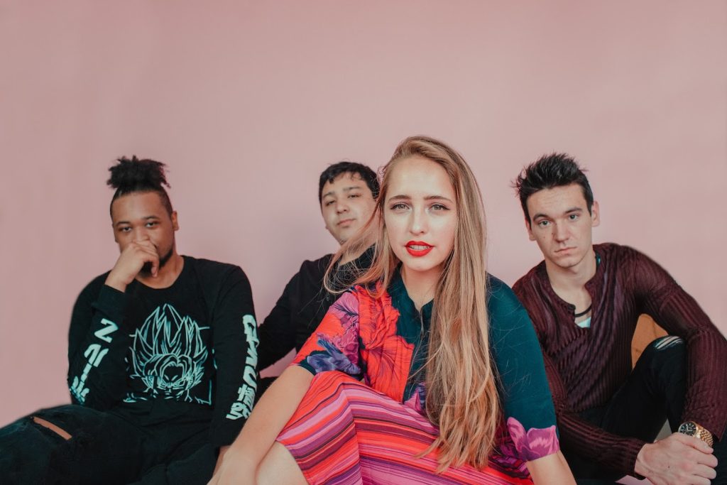 Alex and her bandmates sit in front of a pink wall looking into the camera with a mix of expressions.