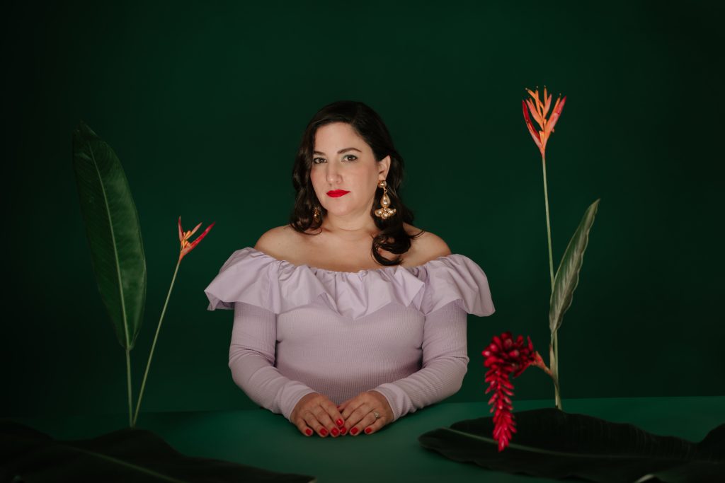 Ani Cordero sits behind a large green table and a green backdrop wearing a frilly pink dress and looking calmly into the camera. Her hands rest on the table along with a few plants.