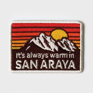 Album art for It's Always Warm In San Araya featuring a sewn patch with the song's title beneath a set of mountains in front of a sunset.