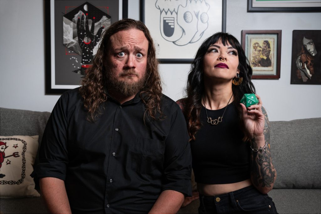 A band photo of the duo on a couch with framed pictures behind them.