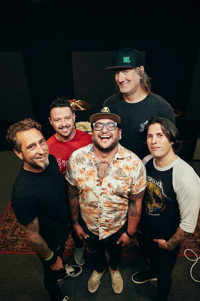 5 bandmates are gathered and smiling towards the camera while standing in front of a drum kit against a black backdrop.