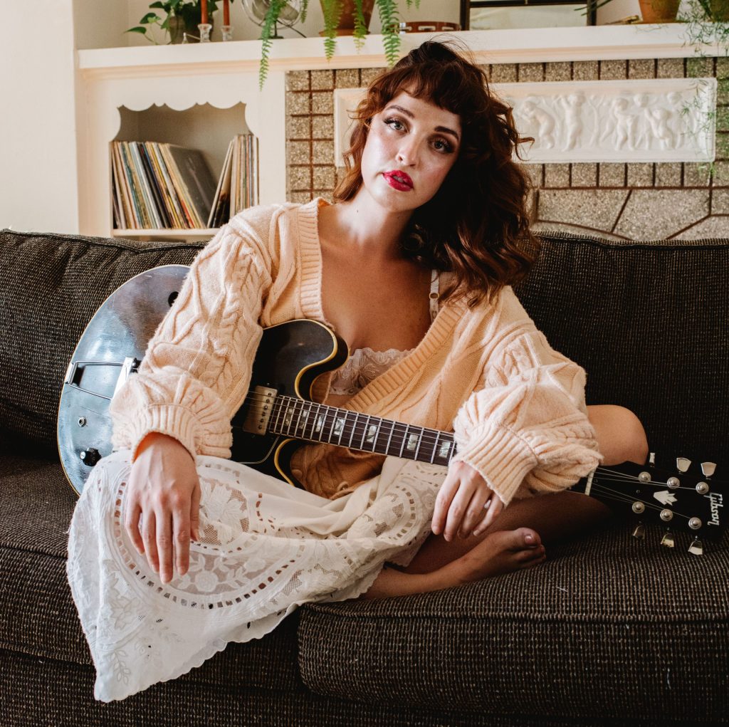 Elizabeth Moen sits on a couch holding an electric guitar.