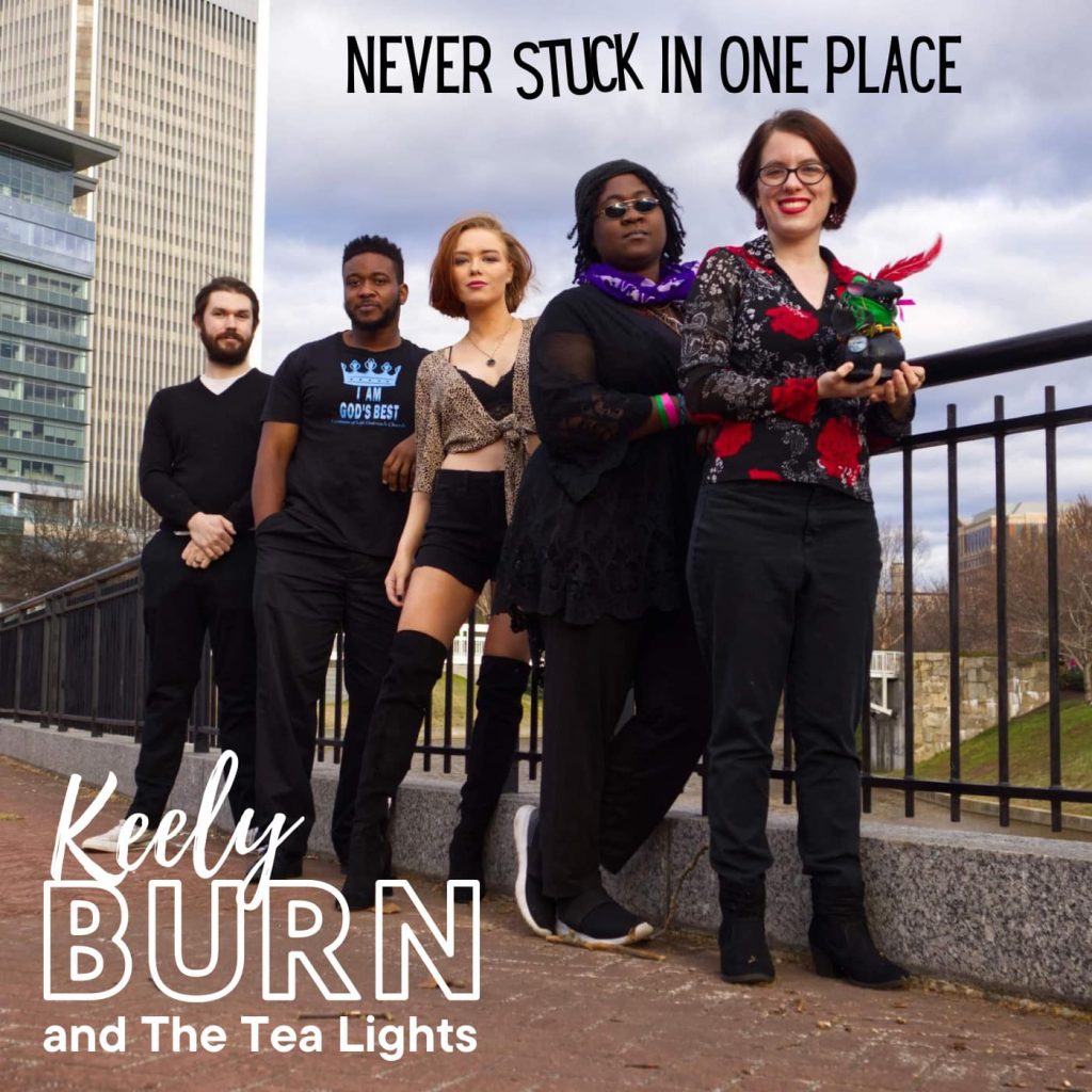 The official album art for Never Stuck In One Place featuring the 5 members of the band lined up on a sidewalk against a railing. Behind them are a few building, a bridge, and an open sky.