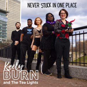 The official album art for Never Stuck In One Place featuring the 5 members of the band lined up on a sidewalk against a railing. Behind them are a few building, a bridge, and an open sky.