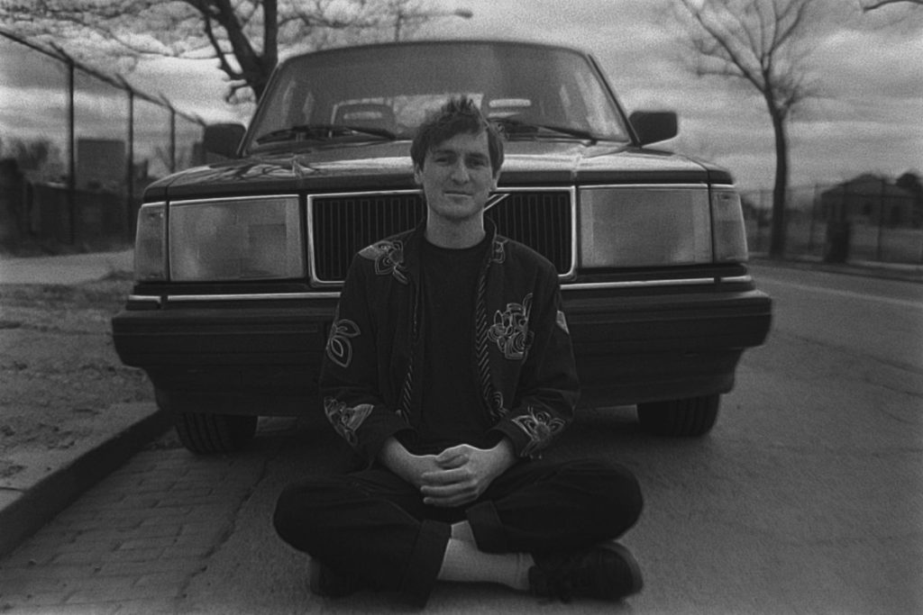 A black and white photo of Poppy Patica's Peter Hartmann sitting in front of a car criss-cross on the road.