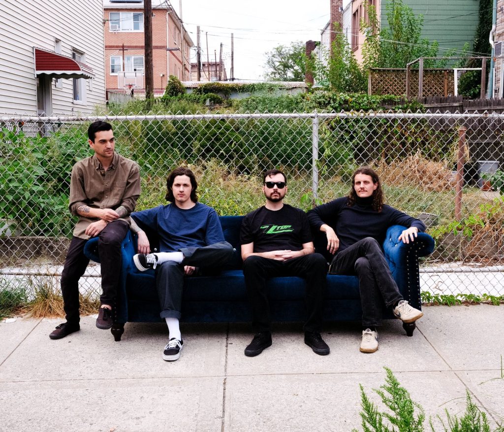 4 bandmates of Plight sit on a blue velvet couch placed on an outdoor sidewalk in front of a chain fence and overgrown backyard in a city setting.