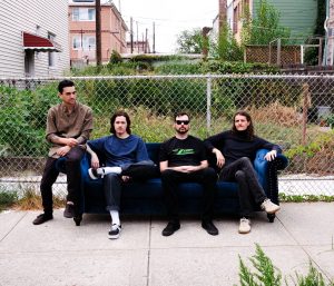 4 bandmates of Plight sit on a blue velvet couch placed on an outdoor sidewalk in front of a chain fence and overgrown backyard in a city setting.