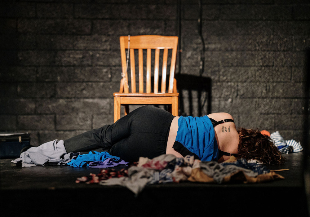 A woman in a blue tank top and black leggings is laying on her side in front of a wooden chair, in the middle of a spotlight. Her back is to the viewer. She is surrounded by dirty clothes.