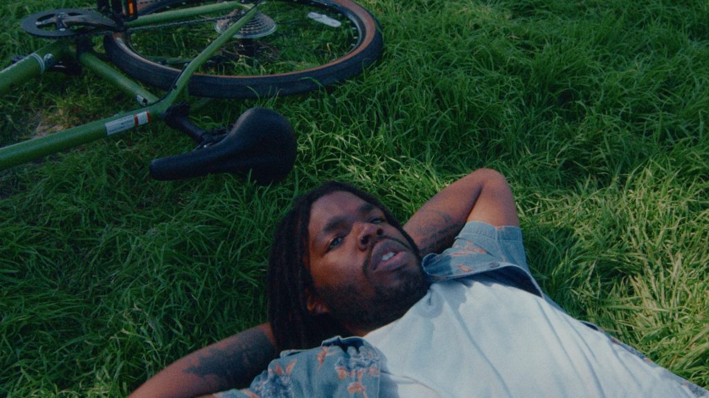 Still of JayWood in music video, laying on grass looking up at sky next to bike