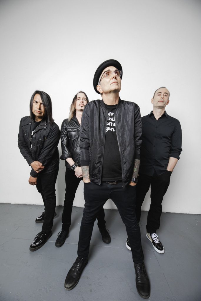 Members of Alternative Rock band Everclear, wearing black while standing on a great concrete floor along a painted white wall.