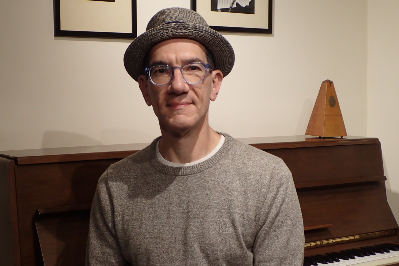 Oren Levine, wearing a hat, sits in front of an upright piano, on which rests a metronome.