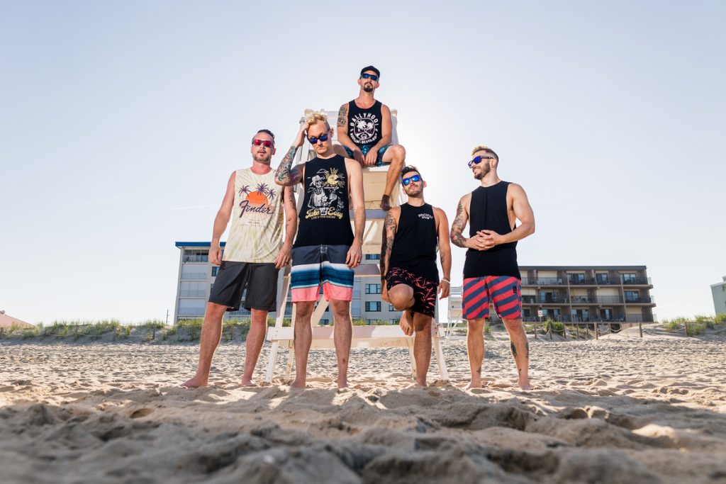 Ballyhoo! band members stand on a sandy beach with beach condos in the background.
