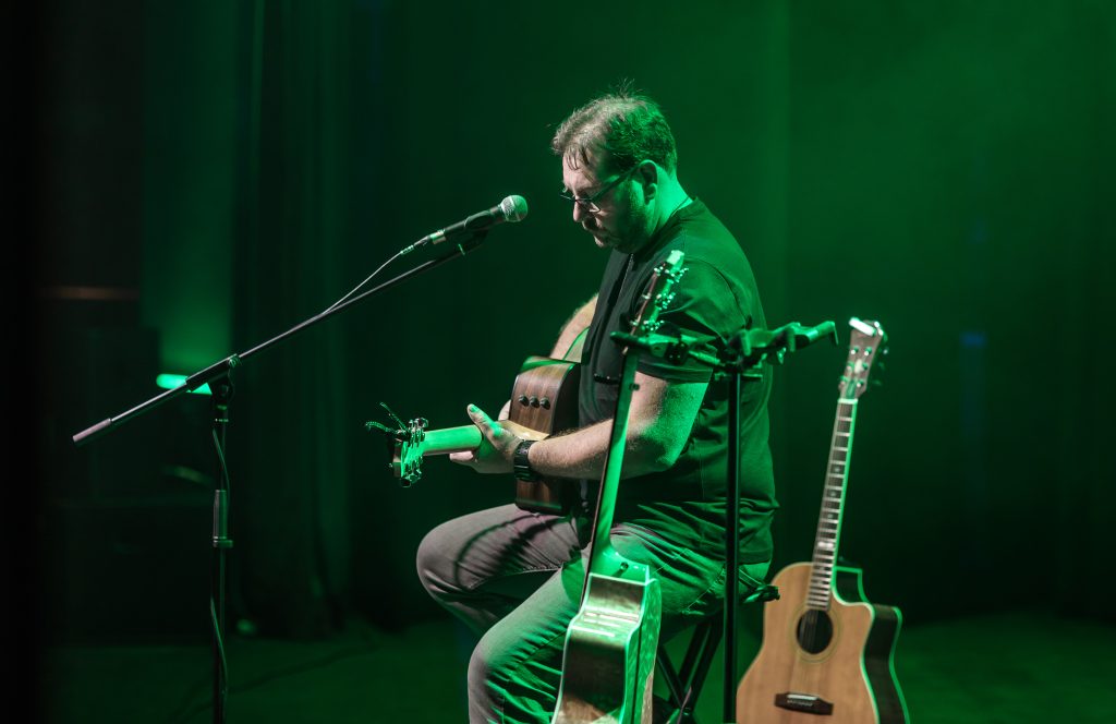 A photo of John Lynch performing on stage, holding a guitar and sitting on a stool. He is lit by green lights.