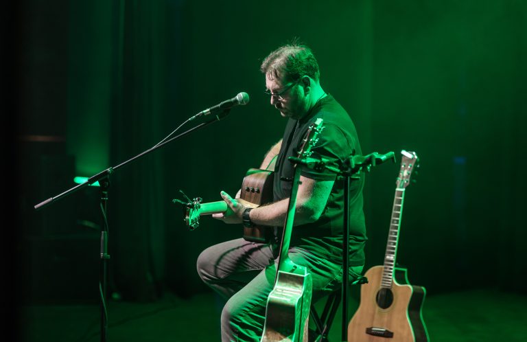 A photo of John Lynch performing on stage, holding a guitar and sitting on a stool. He is lit by green lights.