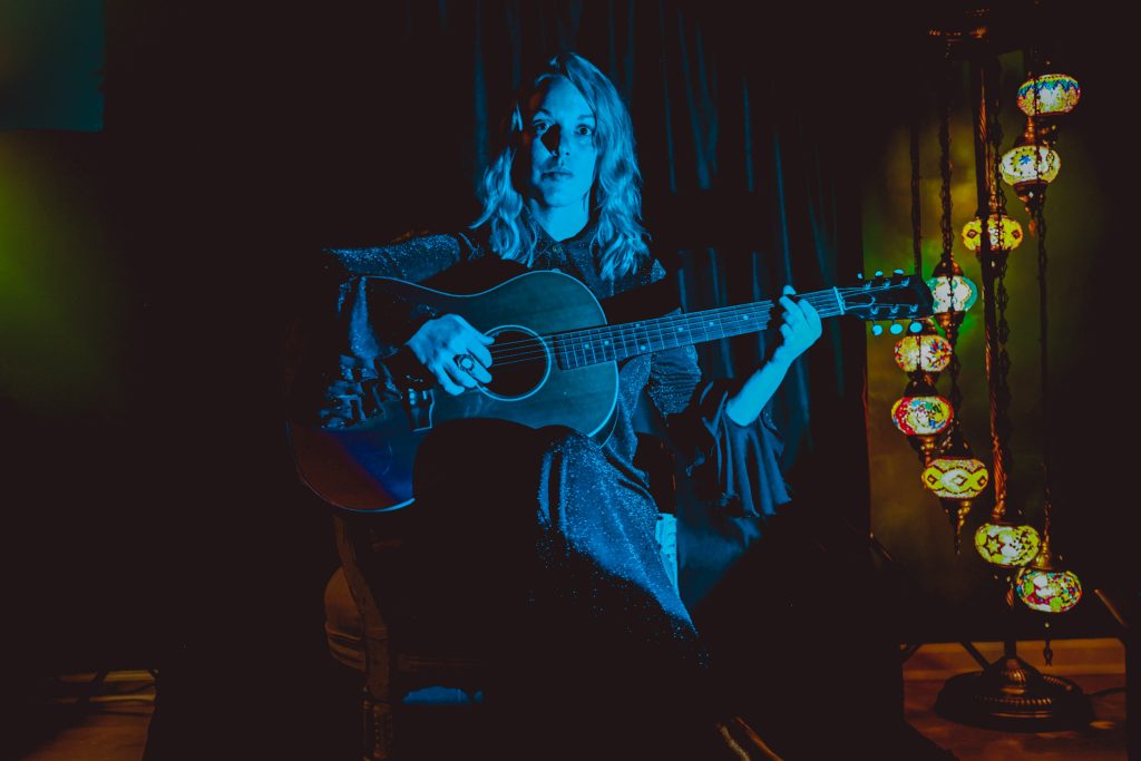Lauren Calve poses for a press photo with an acoustic guitar under blue lighting.