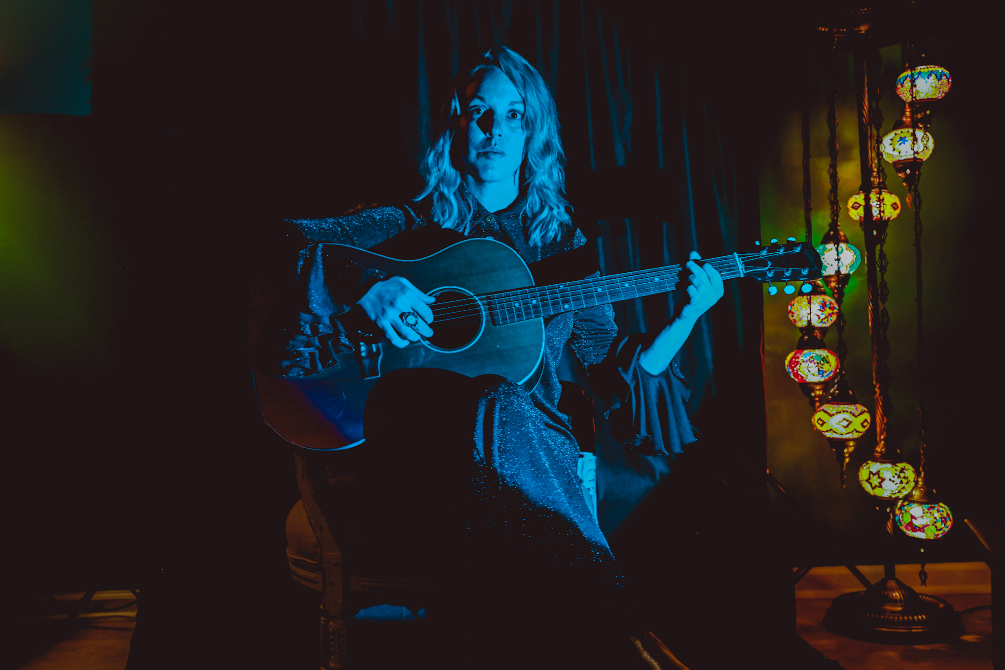 Lauren Calve poses for a press photo with an acoustic guitar under blue lighting.