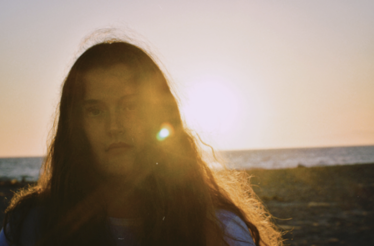 Maggie Cubillos, a young, brunette woman, is standing on a beach and looking at the camera at sunset.