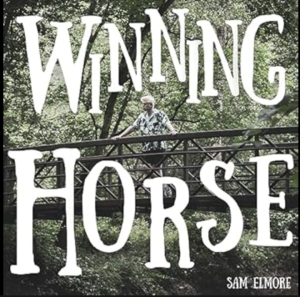 Sam Elmore stands on a wooden bridge surrounded by green foliage.