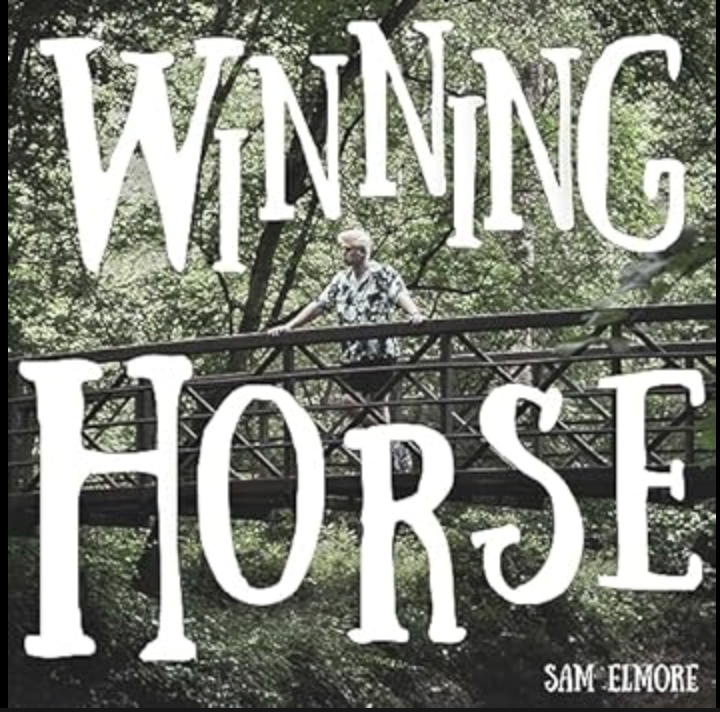 Sam Elmore stands on a wooden bridge surrounded by green foliage.