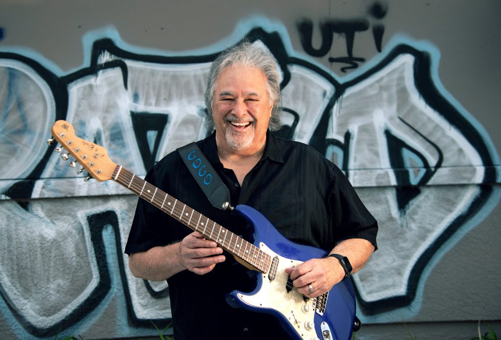 Blues artist Coco Montoya poses with guitar in front of graffiti wall.