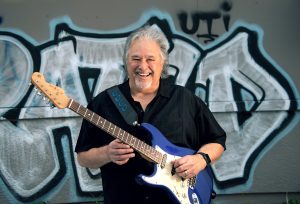 Blues artist Coco Montoya poses with guitar in front of graffiti wall.