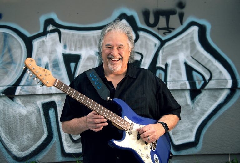 Blues artist Coco Montoya poses with guitar in front of graffiti wall.