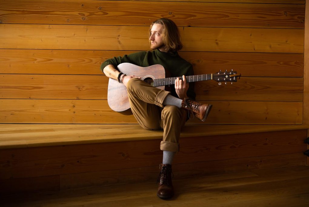 A photo of indie-pop musician Shane Rennison sitting with his guitar inside of a wood panneled room.