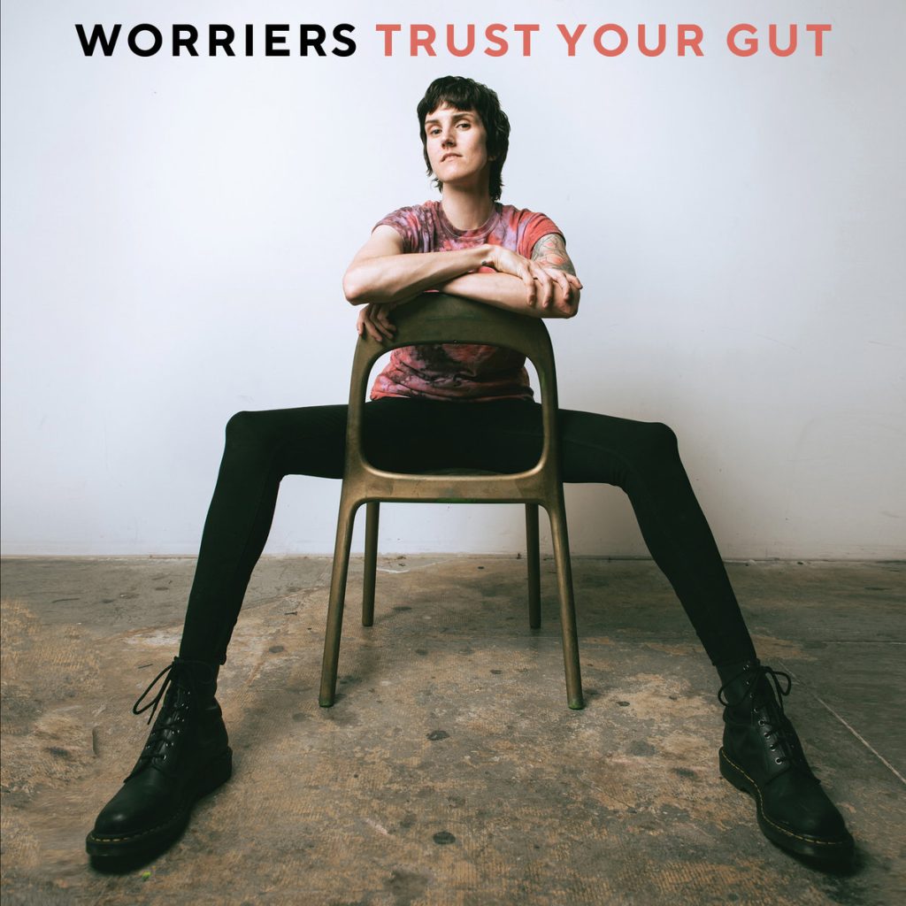 Frontperson of the band, Lauren Denitzo, is sitting with their legs splayed on a backwards wooden chair, resting their arms on the top of the chair. They are in a white room with concrete floors. Worriers is written in black text above their head while Trust Your Gut is written right next to it in red text.