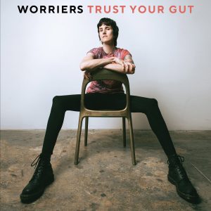 Frontperson of the band, Lauren Denitzo, is sitting with their legs splayed on a backwards wooden chair, resting their arms on the top of the chair. They are in a white room with concrete floors. Worriers is written in black text above their head while Trust Your Gut is written right next to it in red text.