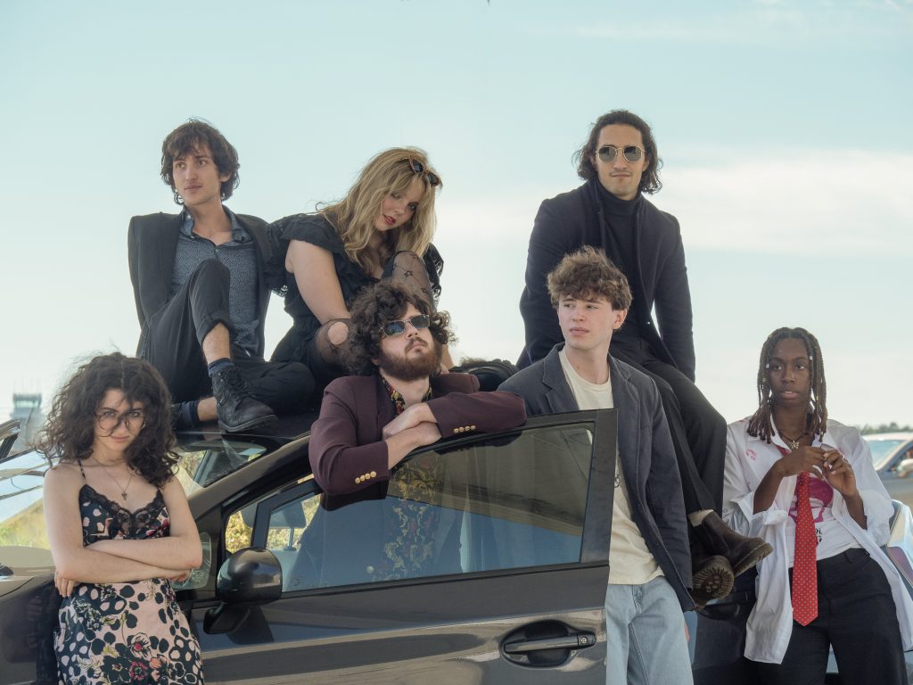 The members of Night Hawk are all leaning against a black car on a windy day, dressed in black.