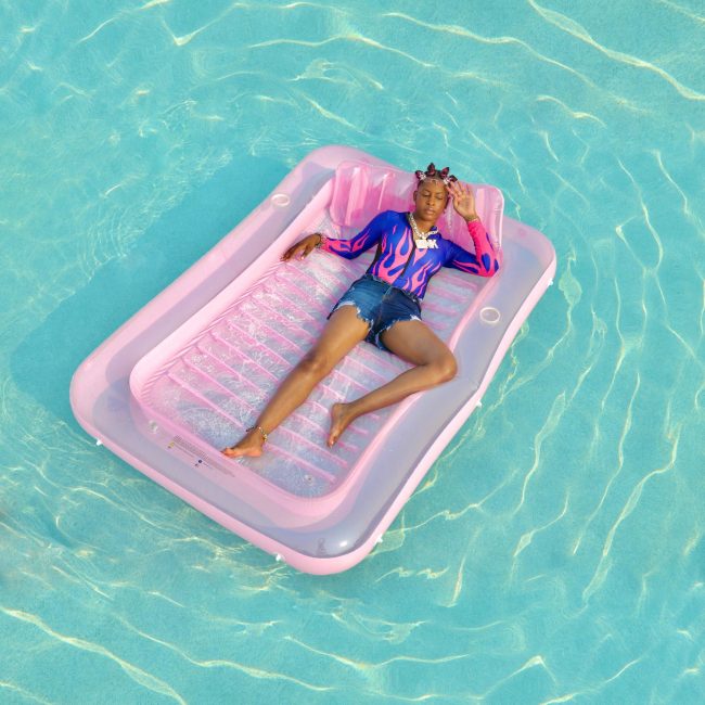 Nia Monaé floats in a pool on a pink raft while wearing jean shorts and a purple nylon swimsuit top with pink flames.