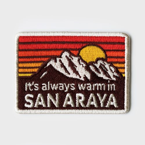 Album art for It's Always Warm In San Araya featuring a sewn patch with the song's title beneath a set of mountains in front of a sunset.
