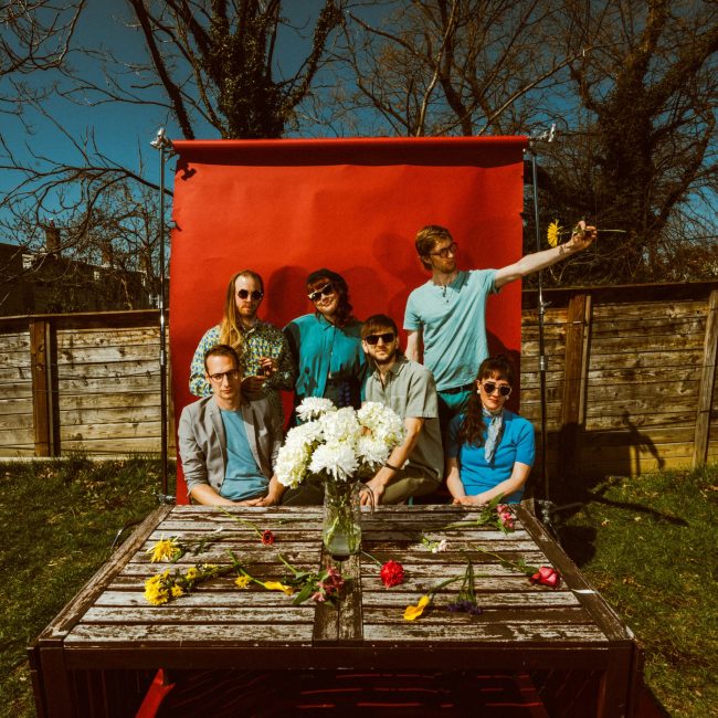The North Country band sits together outside in front of a red photo backdrop. A wooden table in front of the band displays yellow and white flowers.