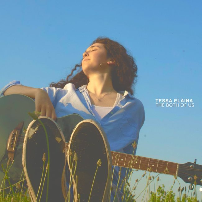Tessa Elaina sitting against a blue sky with her guitar in her lap and long grass in the foreground.