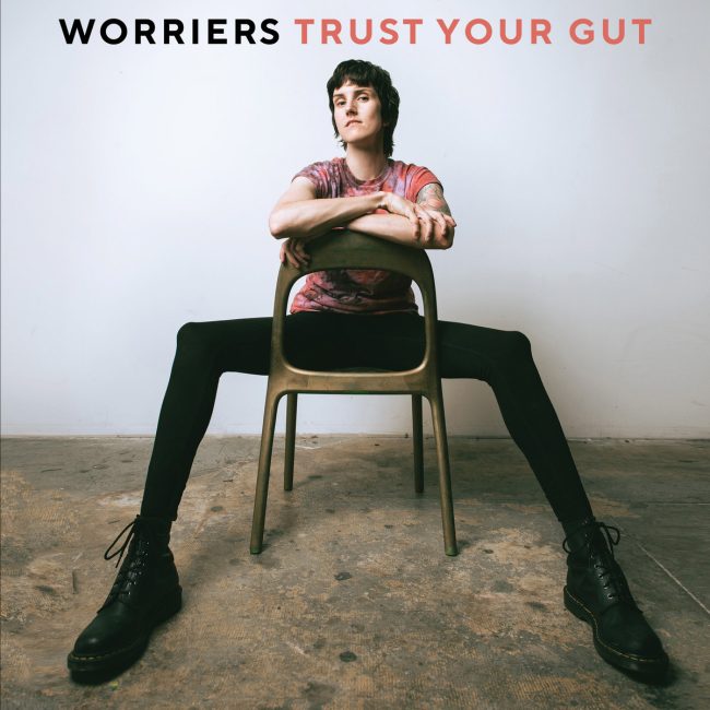 Frontperson of the band, Lauren Denitzo, is sitting with their legs splayed on a backwards wooden chair, resting their arms on the top of the chair. They are in a white room with concrete floors. Worriers is written in black text above their head while Trust Your Gut is written right next to it in red text.